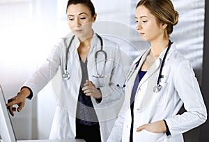 Two female physicians are discussing medical therapy, while standing at the table in a sunny clinic office. Doctors use