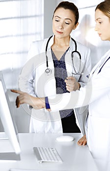 Two female physicians are discussing medical therapy, while standing at the table in a sunny clinic office. Doctors use