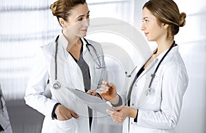 Two female physicians are discussing medical therapy, while standing at the table in a sunny clinic office. Doctors use