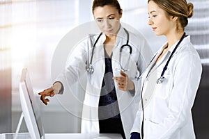 Two female physicians are discussing medical therapy, while standing at the table in a sunny clinic office. Doctors use