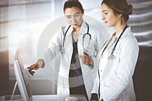 Two female physicians are discussing medical therapy, while standing at the table in a sunny clinic office. Doctors use