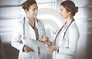 Two female physicians are discussing medical therapy, while standing at the table in a sunny clinic office. Doctors use