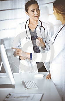 Two female physicians are discussing medical therapy, while standing at the table in a sunny clinic office. Doctors use