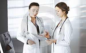Two female physicians are discussing their patient`s medical tests, while standing in a clinic office. Doctors use a