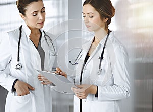 Two female physicians are discussing their patient`s medical tests, while standing in a sunny clinic office. Doctors use