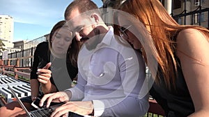 Two female students and a male student communicate on a bench in sunny weather using a laptop and smartphone