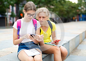 Two female students use phones to check lessons and write down completed assignments in notebook