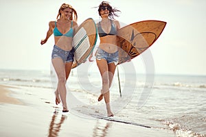 Two female surfers enjoying running on the beach holding surfboards, laughing