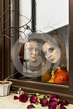 Two female witches look through a spider web ominously out of the window against the backdrop of Halloween decorations. Masquerade