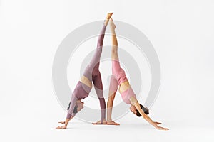 Two female yoga practitioners doing a variation of the Adho Mukha Svanasana exercise on a white background