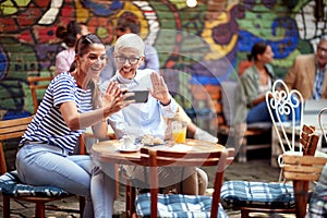 Two females, different ages, sitting in outdoor cafe, using cell phone for online communication, talking to somebody, waving