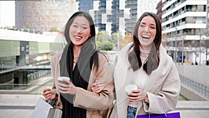 Two femenine multiracial friends walking and talking with coffee cups and shopping bags. A couple of beauty young women