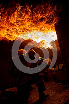 Two firefighters in a burning house full of flames while trying to put out the fire. The men wear respiratory protection equipment