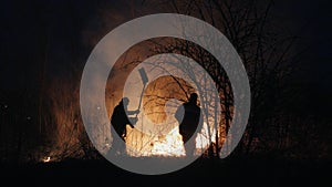 Two firefighters with fire flappers extinguish a fire in the forest at night