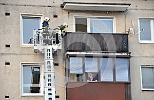 Two firefighters into telescopic boom basket of fire truck try to get to the flat balcony. Old woman is watching them from next ba