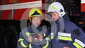 Two firemen in full equipped clothes standing outside at the fire truck with a tablet in hands and deciding what to do