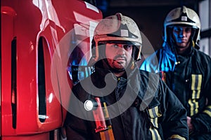 Two firemen wearing protective uniform standing next to a fire truck in a garage of a fire department.
