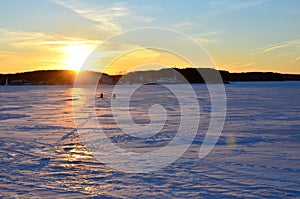 Two fishermen on a frozen lake catch fish in a hole of ice on the background of evening sunset. Winter arrives, bodies of water