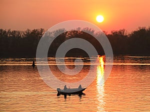 Two fishermen, a man and a woman, swim in a motor boat on the river against the background of an orange sunset. Romantic