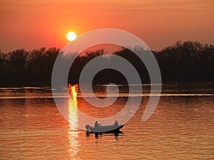 Two fishermen, a man and a woman, swim in a motor boat on the river against the background of an orange sunset. Romantic