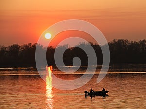 Two fishermen, a man and a woman, swim in a motor boat on the river against the background of an orange sunset. Romantic