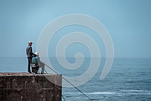 Two fishermen sitting on a pier in front of the water on a gloomy day