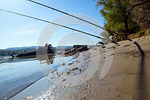 Two fishing rods are mounted on a rack located on a river sandy beach. The day is beautiful and sunny and the angler is waiting