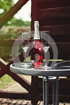 Two flute glasses and an pink champagne bottle with no label on a glass table in a back yard.