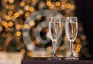 Two flute glasses with sparkling drink champagne standing on dark wooden table. Blurred decorated Christmas fir tree behind