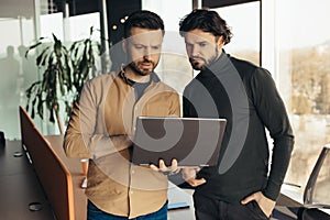 Two focused male employees developing business strategies, cooperating with each other, using laptop pc at modern office