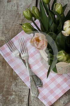 Two forks lie on pink napkin next to a vase of delicate pink tulips. The composition stands on a wooden old background