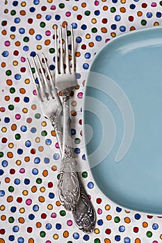 Two forks lie on a tablecloth next to blue plate