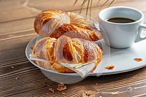 Two French croissants with ham and cheese and a cup of black coffee on the white plate. Wooden table, morning country breakfast