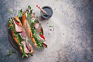 Two fresh baguette sandwiches with meat, tomato, cucumber and arugula on gray background, top view