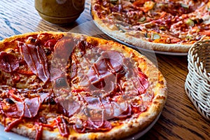 Two fresh baked homemade sliced pizzas with cheese and ham topping served with kitchenware on wooden background.
