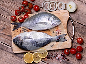 Two fresh gilt-head bream fish on cutting board with lemon, onion and tomato