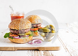 Two fresh homemade burgers, pickles, ketchup and onion rings on white wooden serving board