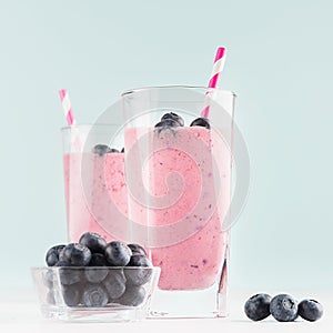 Two fresh purple milkshakes with blueberries in bowl, striped straws closeup on soft light mint color background and white wood.