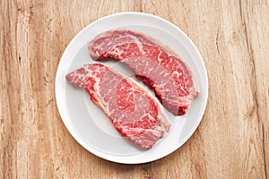 Two fresh raw striploin steak on white plate on wooden background being prepared for cooking