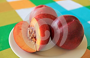 Two Fresh Ripe Nectarine Whole Fruits with One Cut Nectarine on a White Plate Served on Vibrant Color Tablecloth