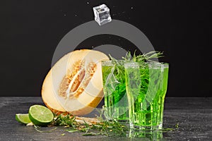Two fresh and summer cold cocktails with ice cubes, tarragon leaves and near a half of a melon on a black background. Refreshing d