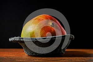 Two fresh whole mango fruit in a black plastic container on a wooden surface,