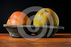 Two fresh whole mango fruit in a black plastic container on a wooden surface