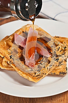 Two, freshly baked, homemade, wild rice, waffles with two slices of ham and warm maple syrup pouring over the meal