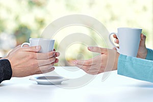 Two friends or couple talking holding coffee cups sitting in a table at home with a window in the