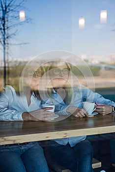 Two friends are enjoying coffee in a coffee shop, looking through a glass with reflections, sitting at a table chatting