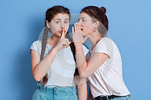 Two friends gossiping isolated over blue background, girl with knot whispering secret and keeps palm near mouth, girl with