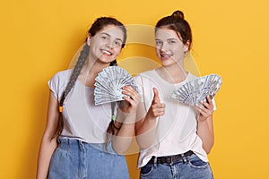 Two friends holding cash in hands, looking at camera with happy facial expression, ladies with dollars, female against yellow wall