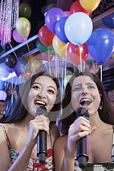 Two friends holding microphones and singing together at karaoke, balloons in the background