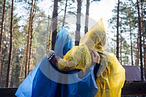 Two friends in hooded raincoats standing back to the camera in a tourist parking lot in the woods. Women in colorful raincoats